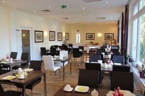 A hotel dining area with multiple tables and chairs, framed artwork on the walls, and large windows providing natural light. - AKZENT Strandresidenz Villa Verdi (Kuehlungsborn)