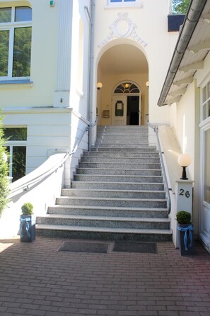 Eine prächtige Treppe führt zu einem Hauseingang, mit einer Nummertafel '26' und einem dekorativen Bogen darüber.