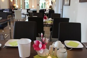 Ein Essbereich mit Tischen für zwei Personen, mit weißen Tellern, Tassen und einer Vase mit rosa Blumen.