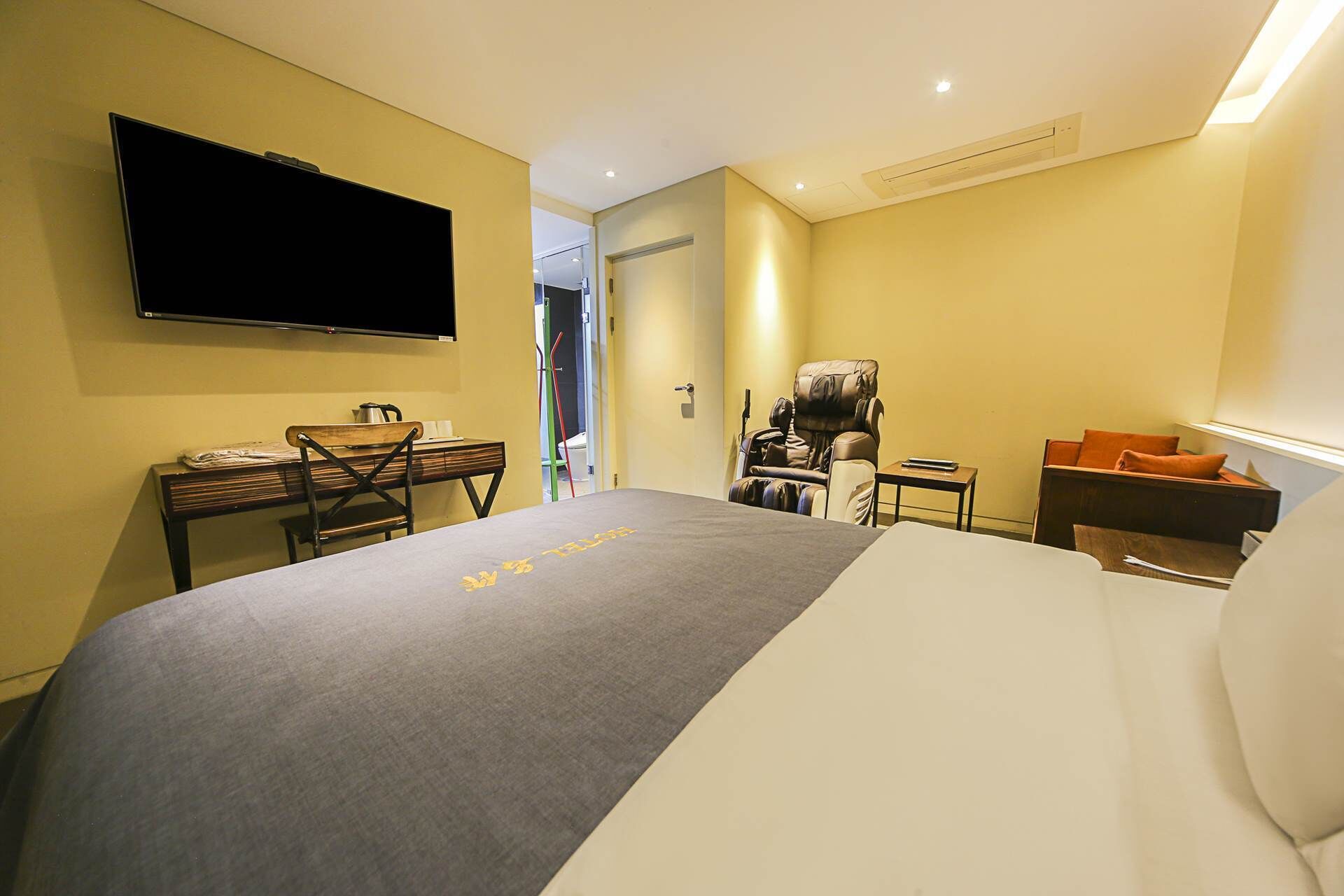 A hotel room with a bed, a desk, a massage chair, and a TV mounted on the wall.
