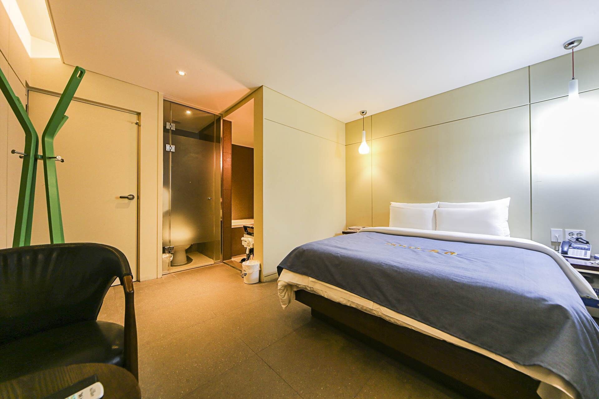 A hotel room with a bed, a chair, a desk with a phone, and a bathroom with a glass door.