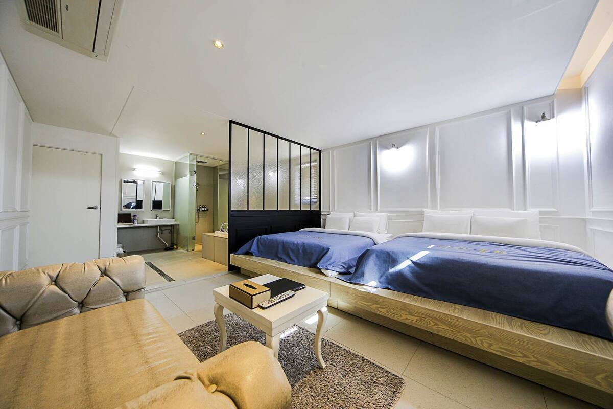 A modern hotel room with a bed, a sofa, a small table, and a bathroom visible through a glass door.