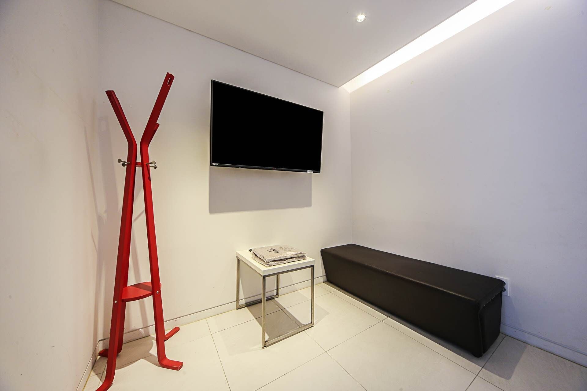 Une pièce au décor minimaliste, avec un téléviseur à écran plat fixé au mur, une chaise en métal rouge, une petite table en verre sur laquelle repose un livre, et un banc noir.