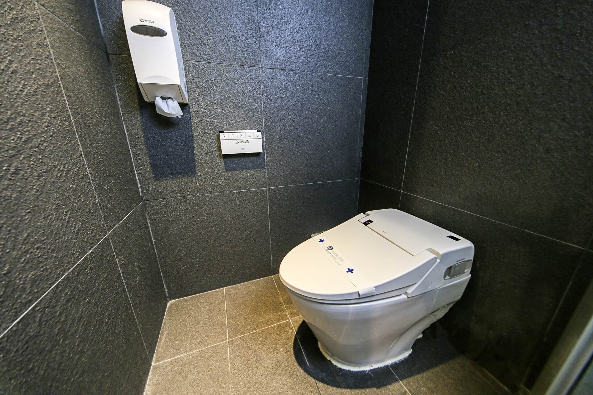 A small, modern bathroom with a toilet, a hand sanitizer dispenser, and a control panel.