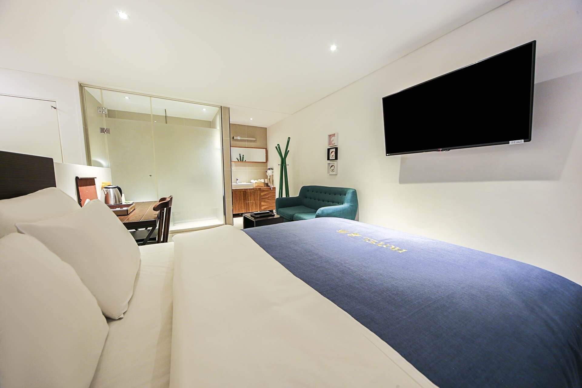 Ein modernes Hotelzimmer mit einem Bett, einem an der Wand befestigten Fernseher, einer Sitzecke mit einem Sessel und einer kleinen Küchenzeile im Hintergrund.