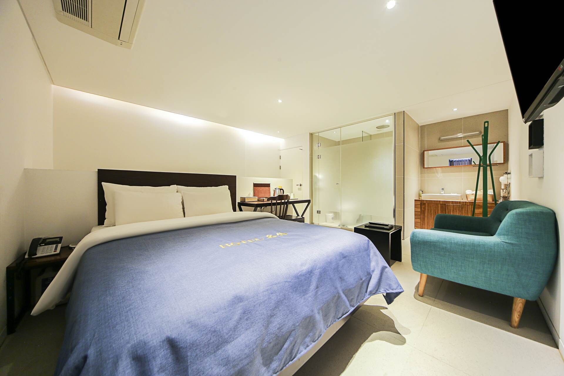 A modern hotel room with a large bed, a desk, a chair, and a glass-enclosed bathroom.