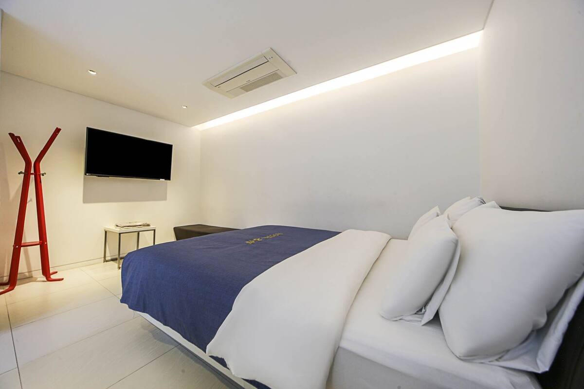 A modern hotel room with a bed, a flat-screen TV, a desk, and a red chair.