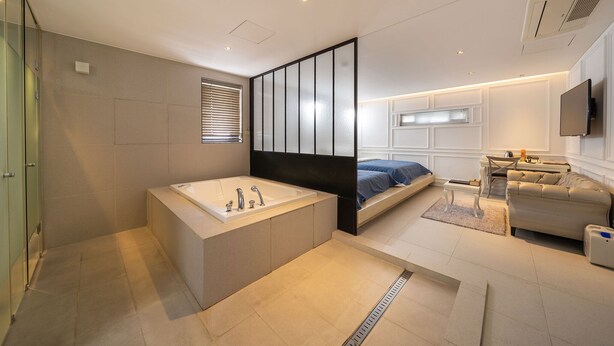 A modern bedroom with a large bathtub, a bed with a blue bedspread, a sofa, and a television mounted on the wall. - Hotel Myeongjak (Chuncheon)