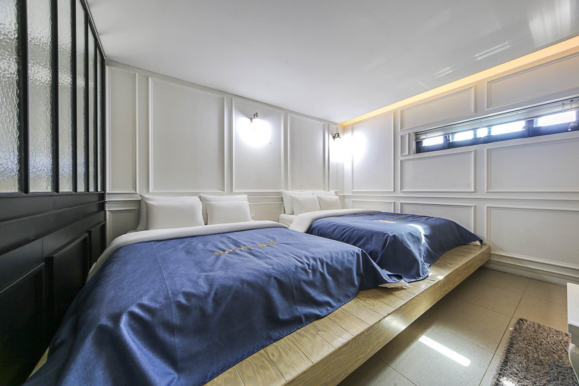 A bedroom with two beds, white walls, and a window with blinds.