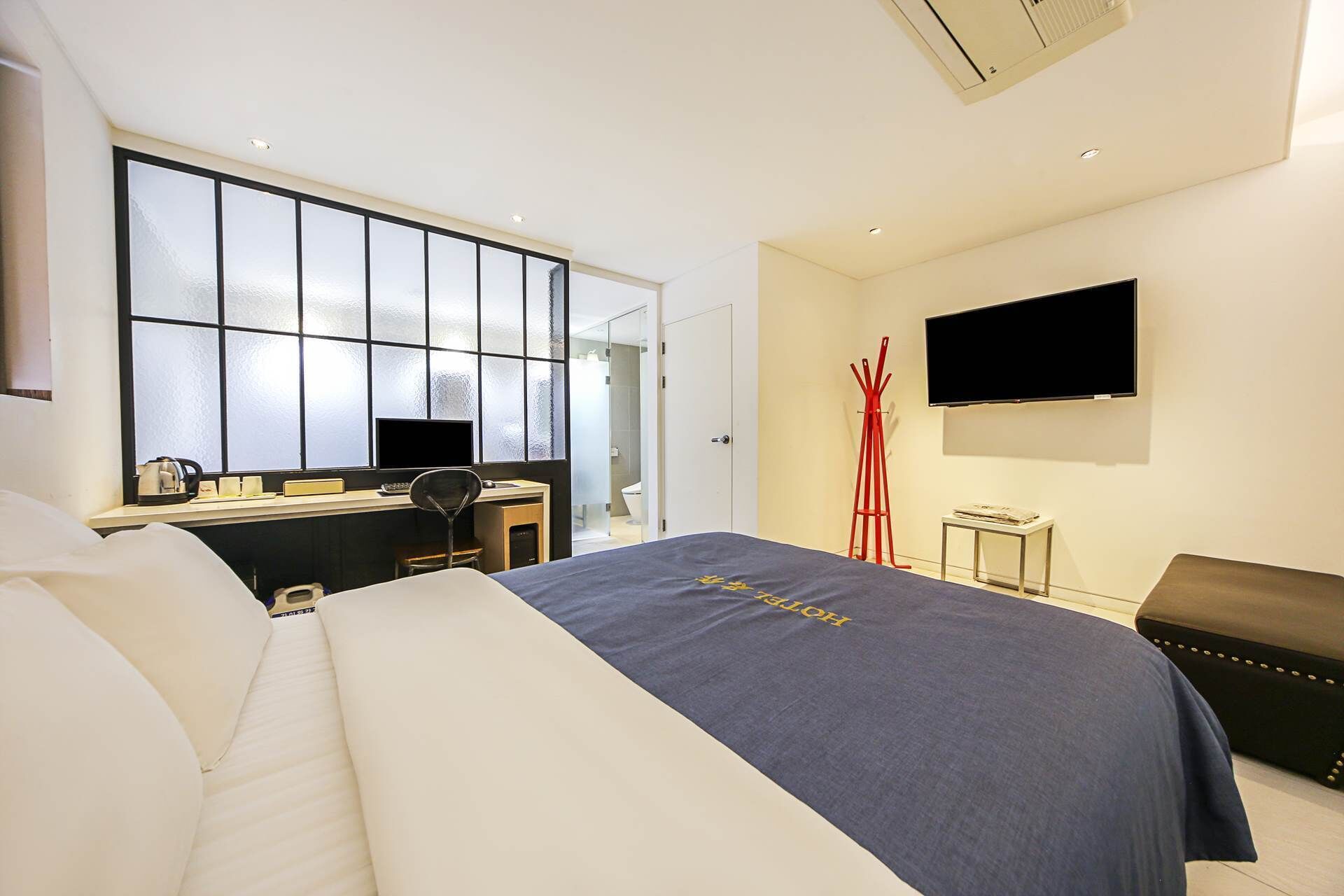 A hotel room with a bed, a desk with a computer, a TV, and a red coat rack.