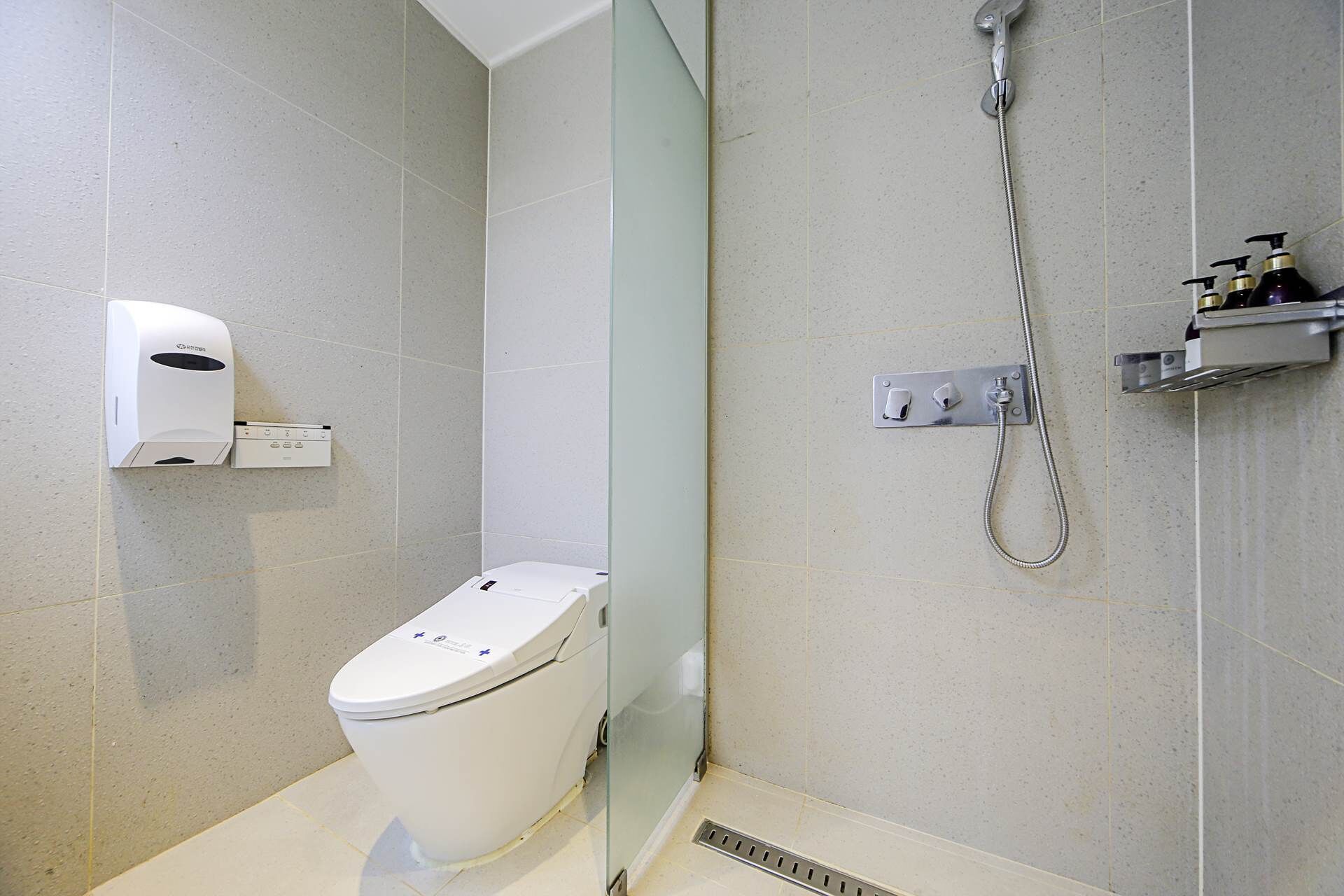 A modern bathroom with a toilet, a hand dryer, a shower with a handheld showerhead, and a soap dispenser.