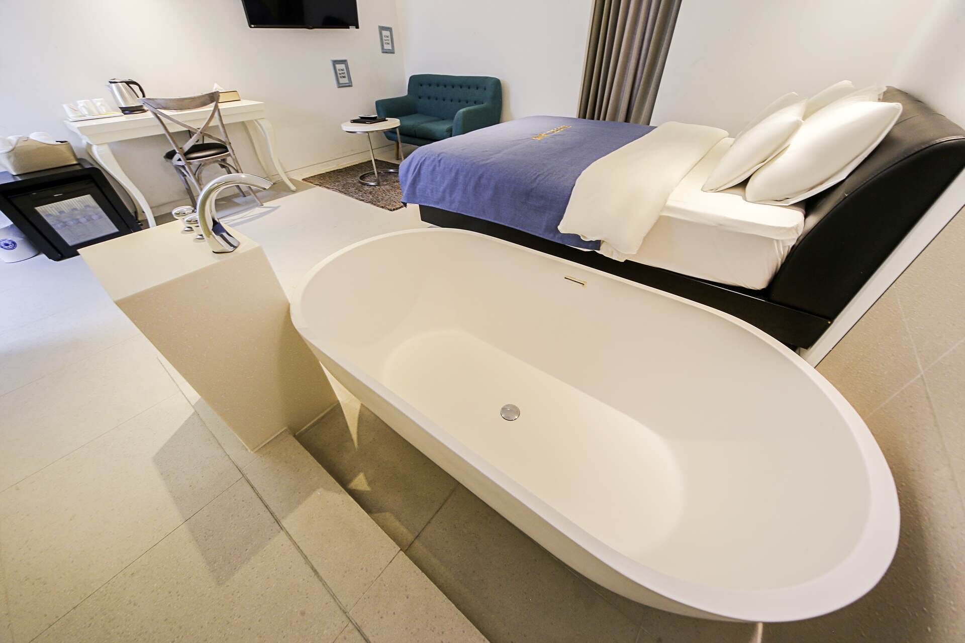 A modern bathroom with a large bathtub, a small bedside table, a chair, and a flat-screen TV mounted on the wall.