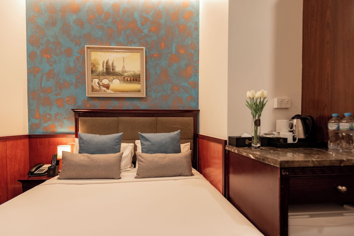 A hotel room with a bed, a nightstand with a phone, a framed picture on the wall, and a small table with a vase of flowers.