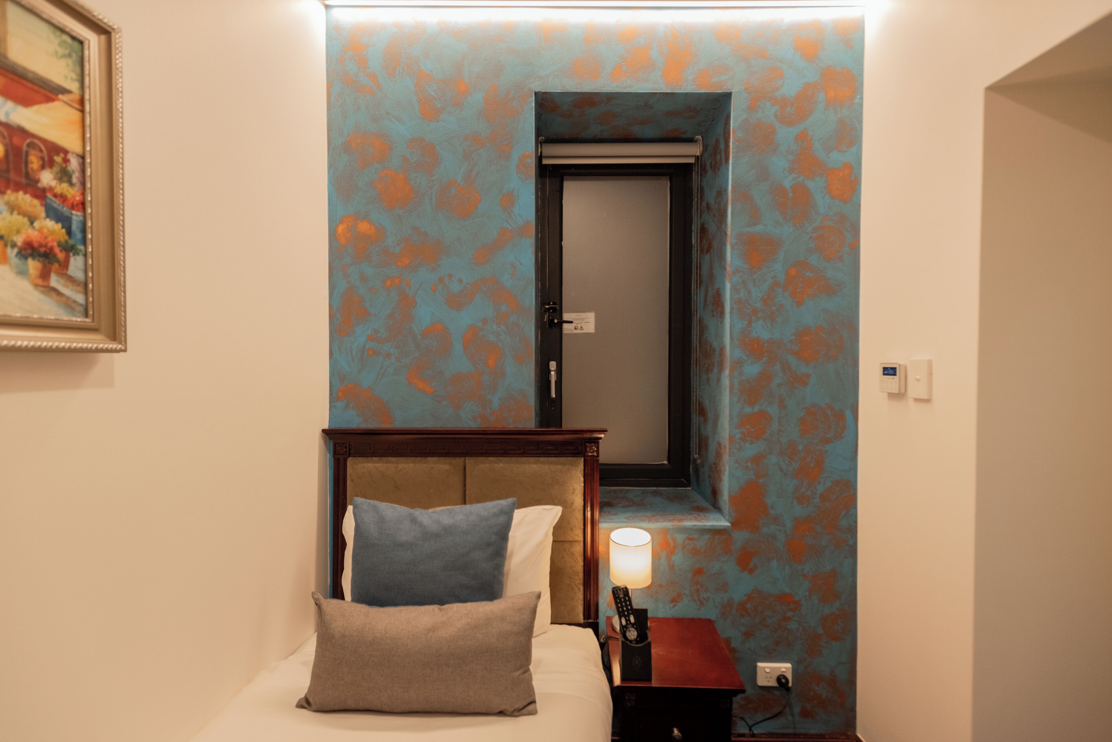 A hotel room with a bed, a nightstand, a lamp, a painting, and a door with a mirror.