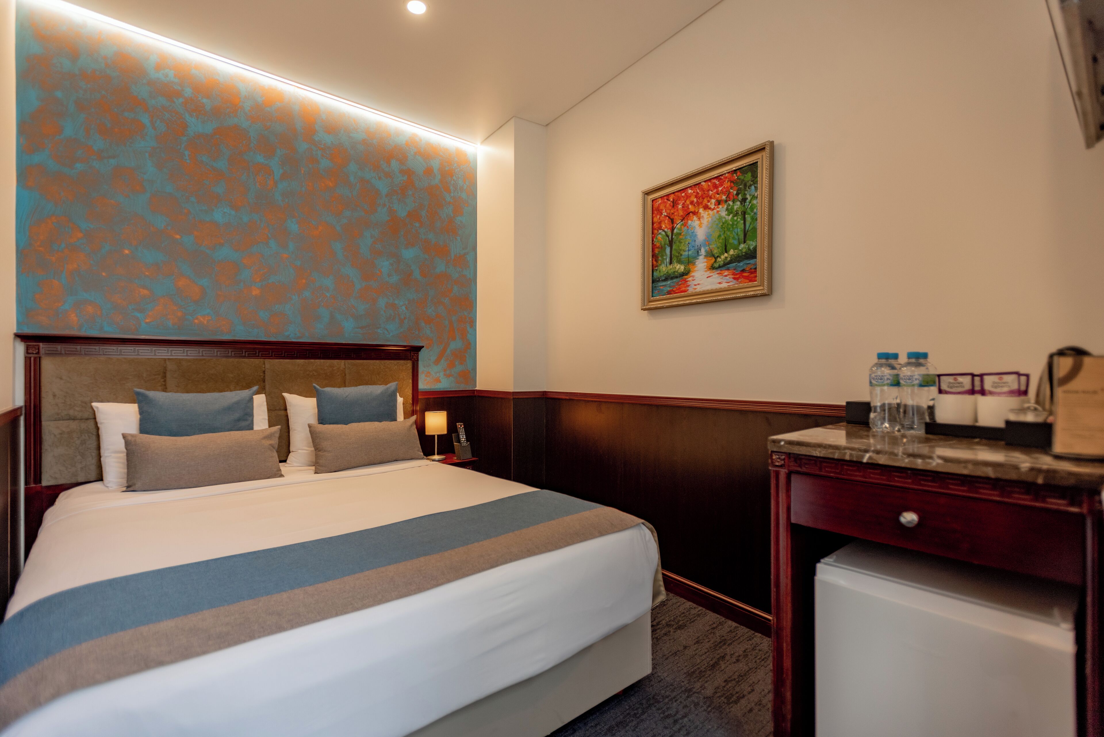 A hotel room with a large bed, a nightstand with a lamp, a small refrigerator, and a painting on the wall.