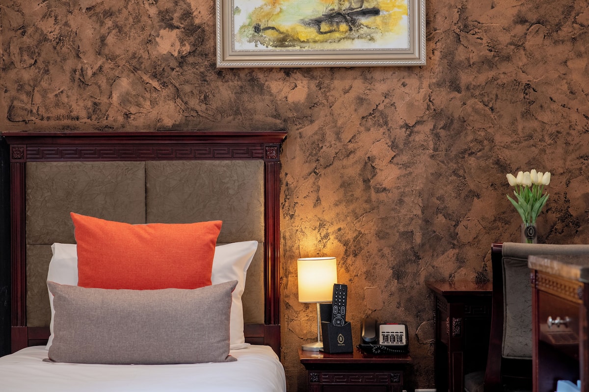 A hotel room with a bed, a nightstand with a lamp, a remote control, and a vase with flowers.
