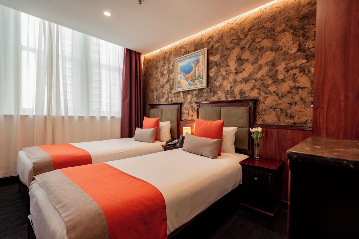 A hotel room with two beds, a nightstand with a phone, a vase with flowers, and a painting on the wall.
