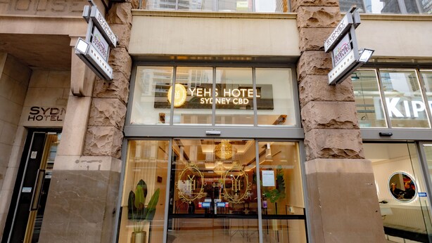 Entrada del Hotel Yeoh’s Sydney CBD con fachada de vidrio moderna, un logotipo dorado destacado y el reflejo de edificios circundantes.