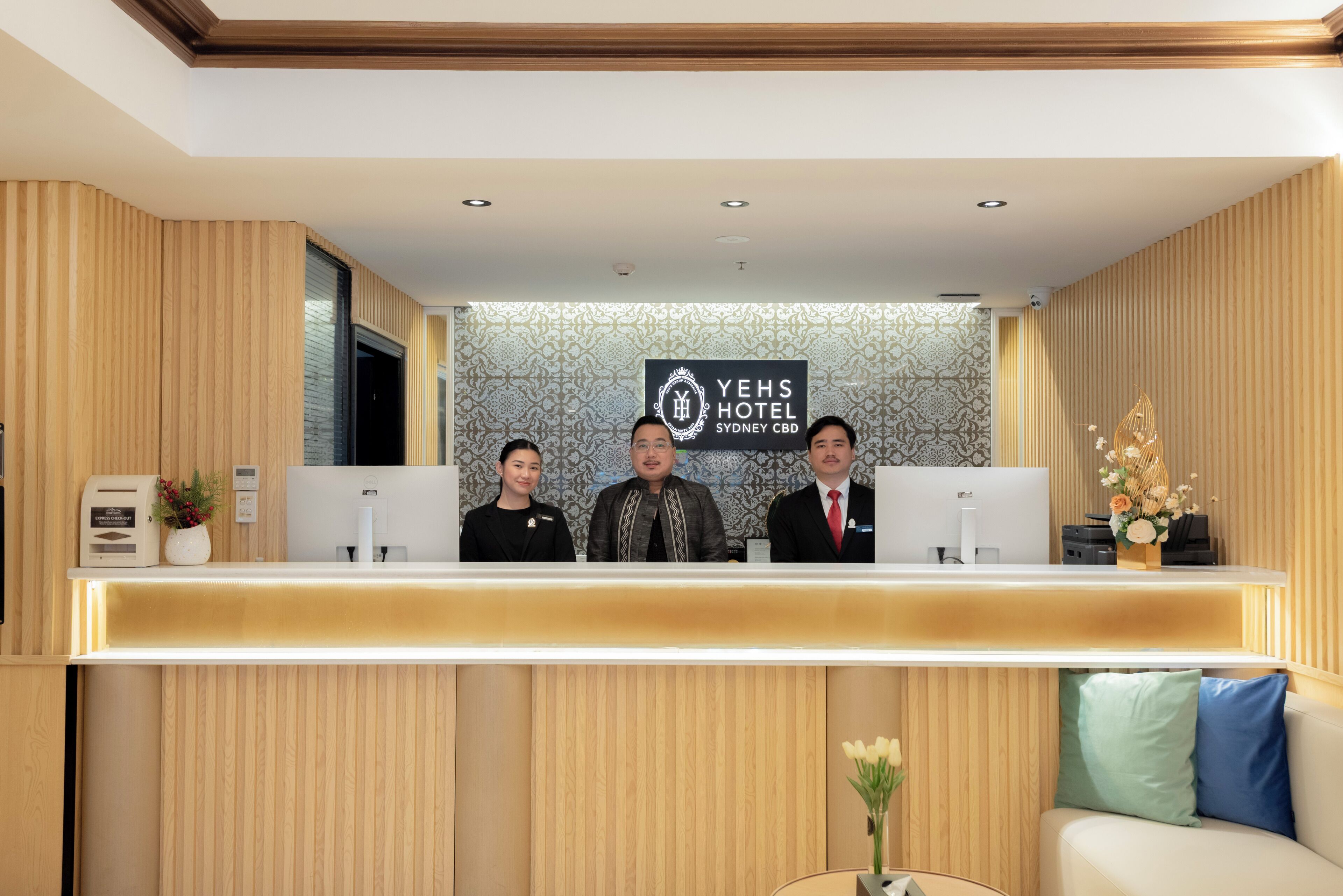 A reception desk at YeHs Hotel Sydney CBD with three staff members, a computer, and a small table with a vase of flowers.