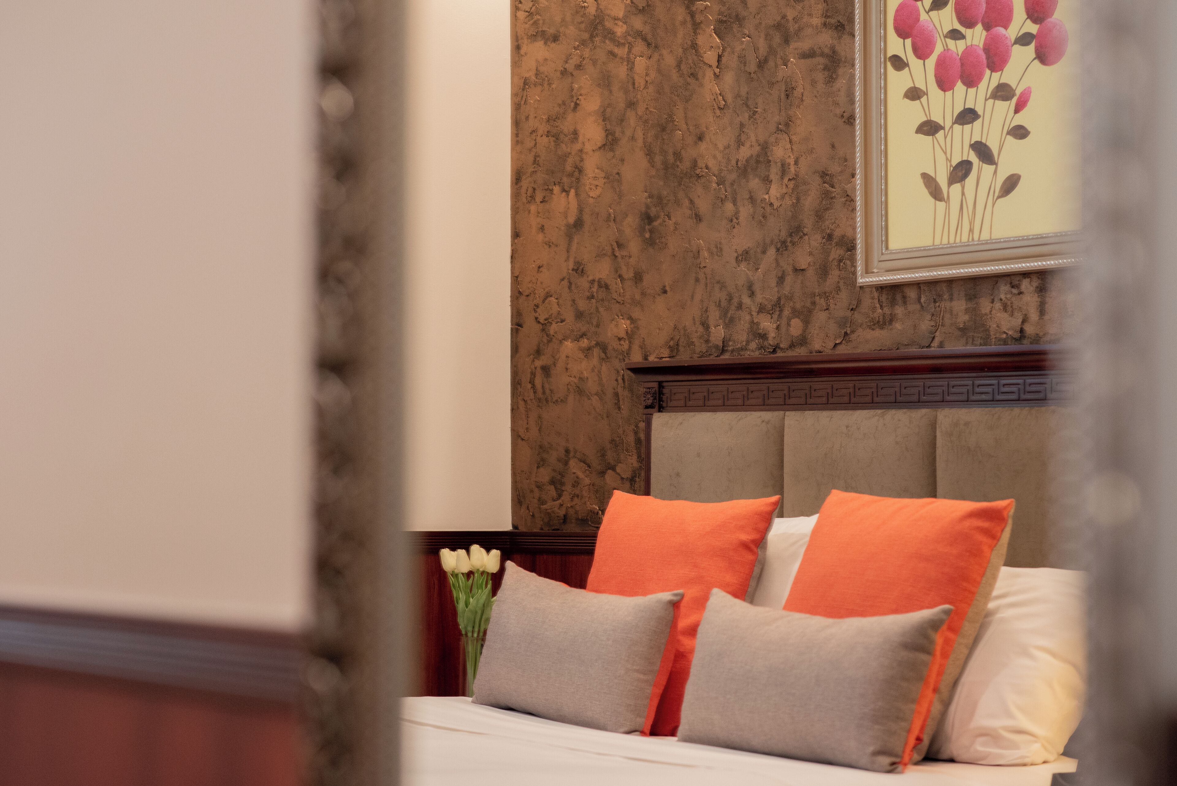 Una cama bien tendida con almohadas grises y naranjas, un cabecero de madera y una obra de arte floral enmarcada en la pared.