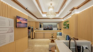 Zona de recepción de un hotel con pared de paneles de madera, una lámpara de araña, mostrador de recepción, televisor y un área de estar con máquina de café.