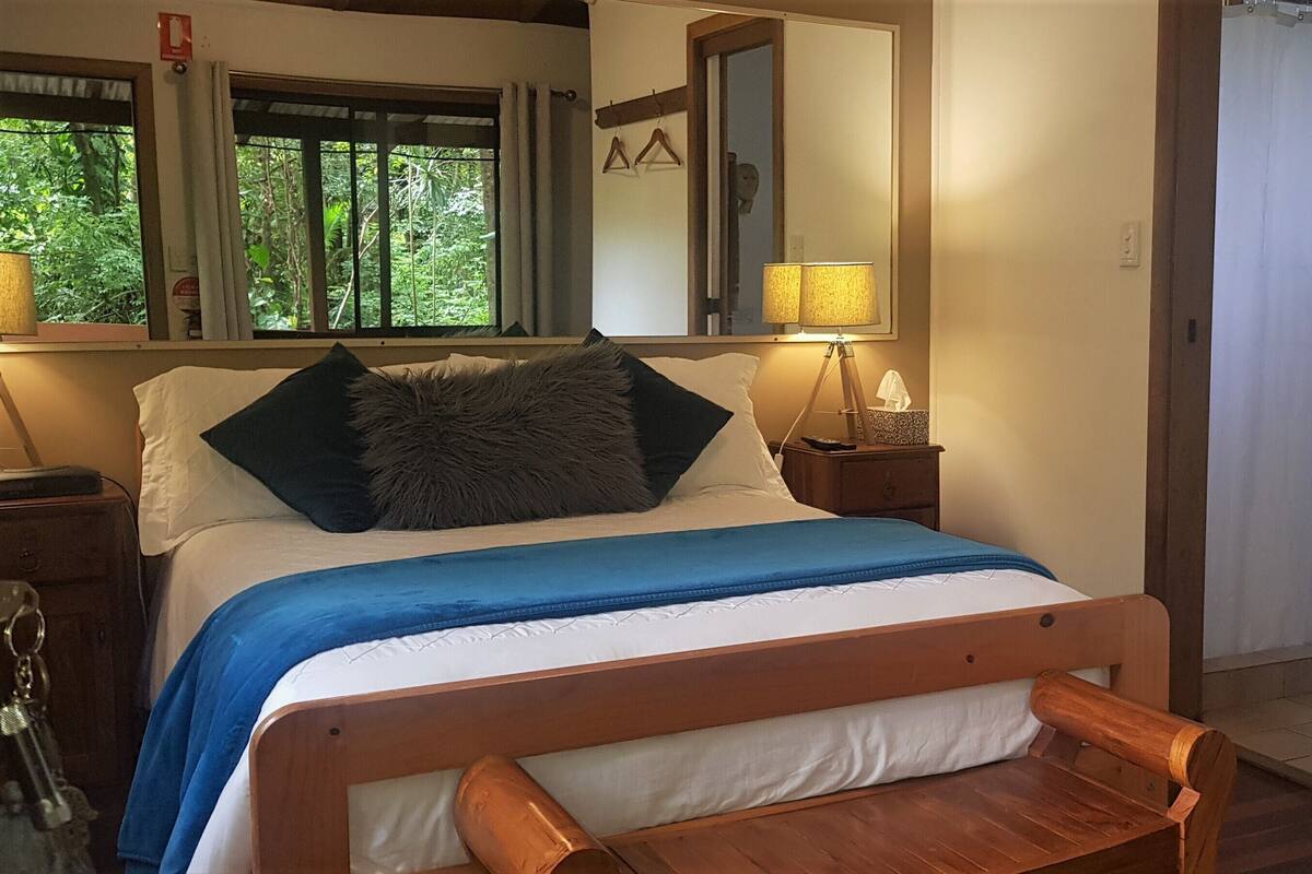 A bedroom with a bed, a wooden bench, a nightstand, a lamp, a mirror, and a window with a view of greenery.