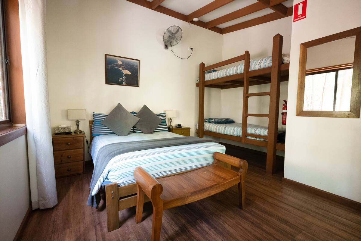 A bedroom with a bunk bed, a wooden bench, a bedside table with a lamp, a framed picture on the wall, and a window with curtains.