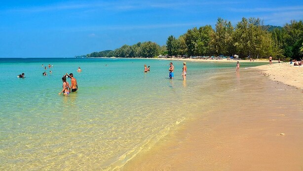 A beach with clear turquoise water, people swimming and walking along the shore, and a line of trees in the background.