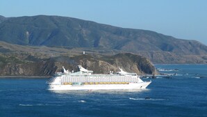 A large cruise ship with multiple decks and a white hull, sailing near a coastline with rocky cliffs and a lighthouse in the background. - Pacific View Bed and Breakfast (Wellington)