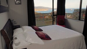 A bedroom with a large bed, a nightstand, a lamp, and a view of the sea and hills. - Pacific View Bed and Breakfast (Wellington)