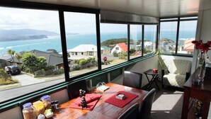 A dining area with a wooden table, chairs, and a view of the ocean and houses. - Pacific View Bed and Breakfast (Wellington)