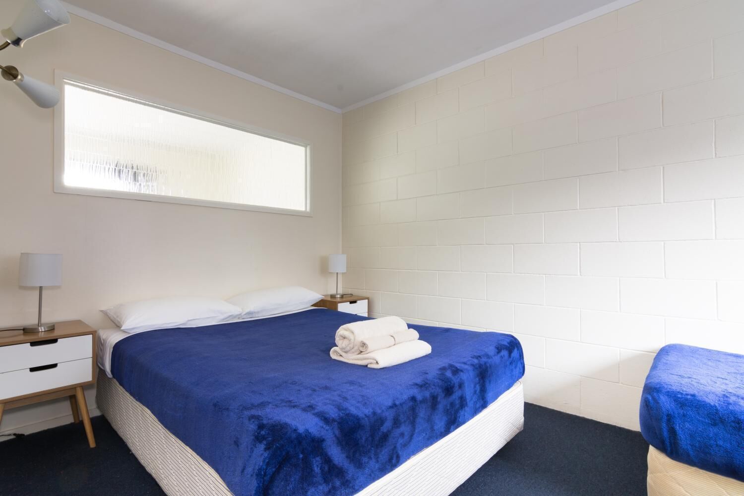 A hotel room with a large bed, two bedside tables, a window with blinds, and a white brick wall.