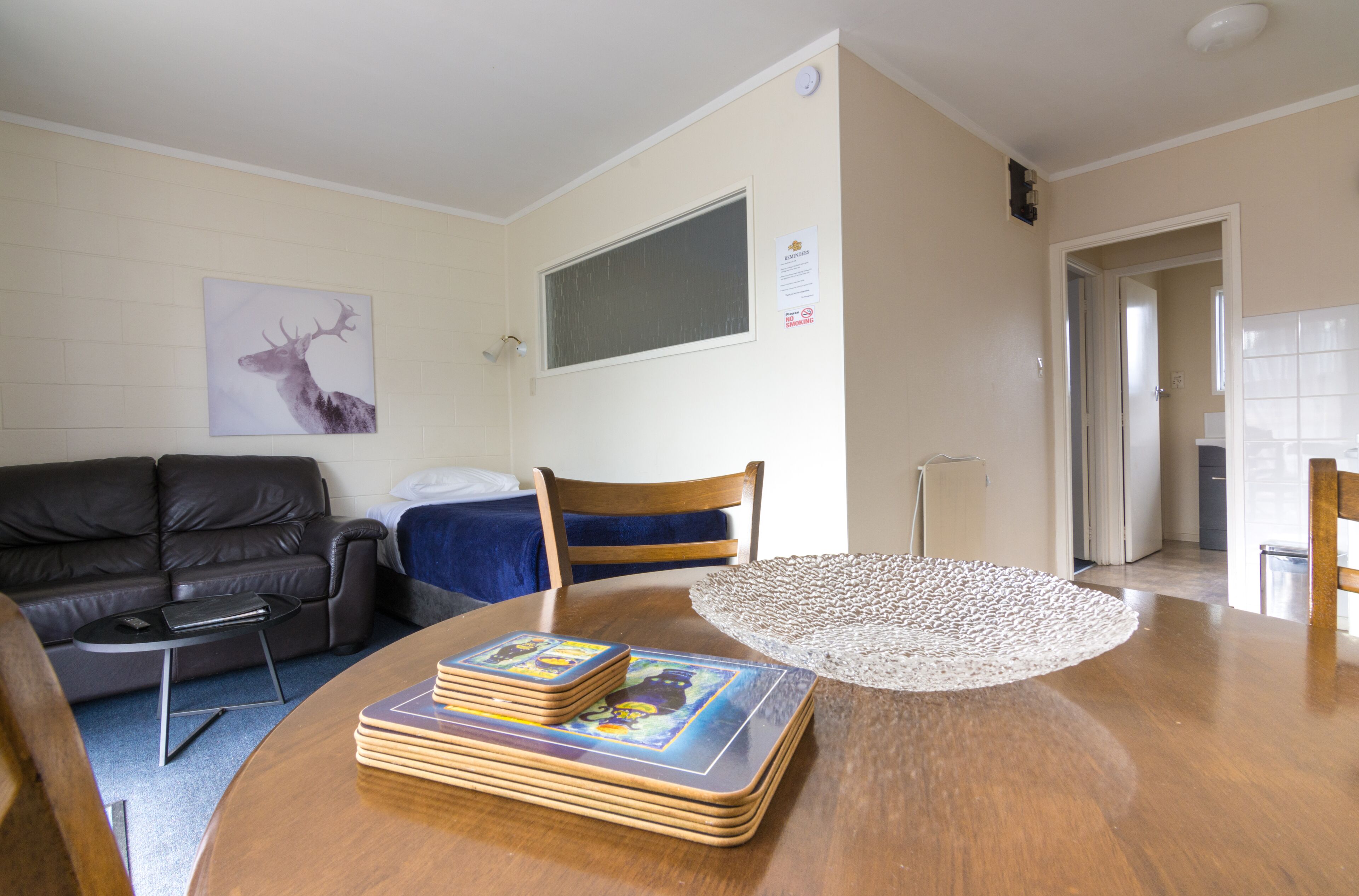 A living room with a wooden table, a black leather sofa, a blue chair, and a painting of a deer on the wall.
