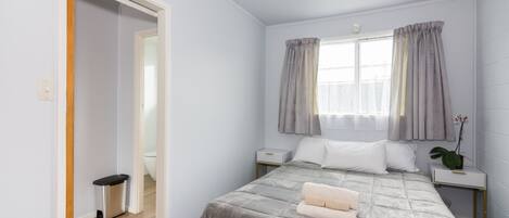A bedroom with a bed, a nightstand, a small trash bin, a window with curtains, and a potted plant.