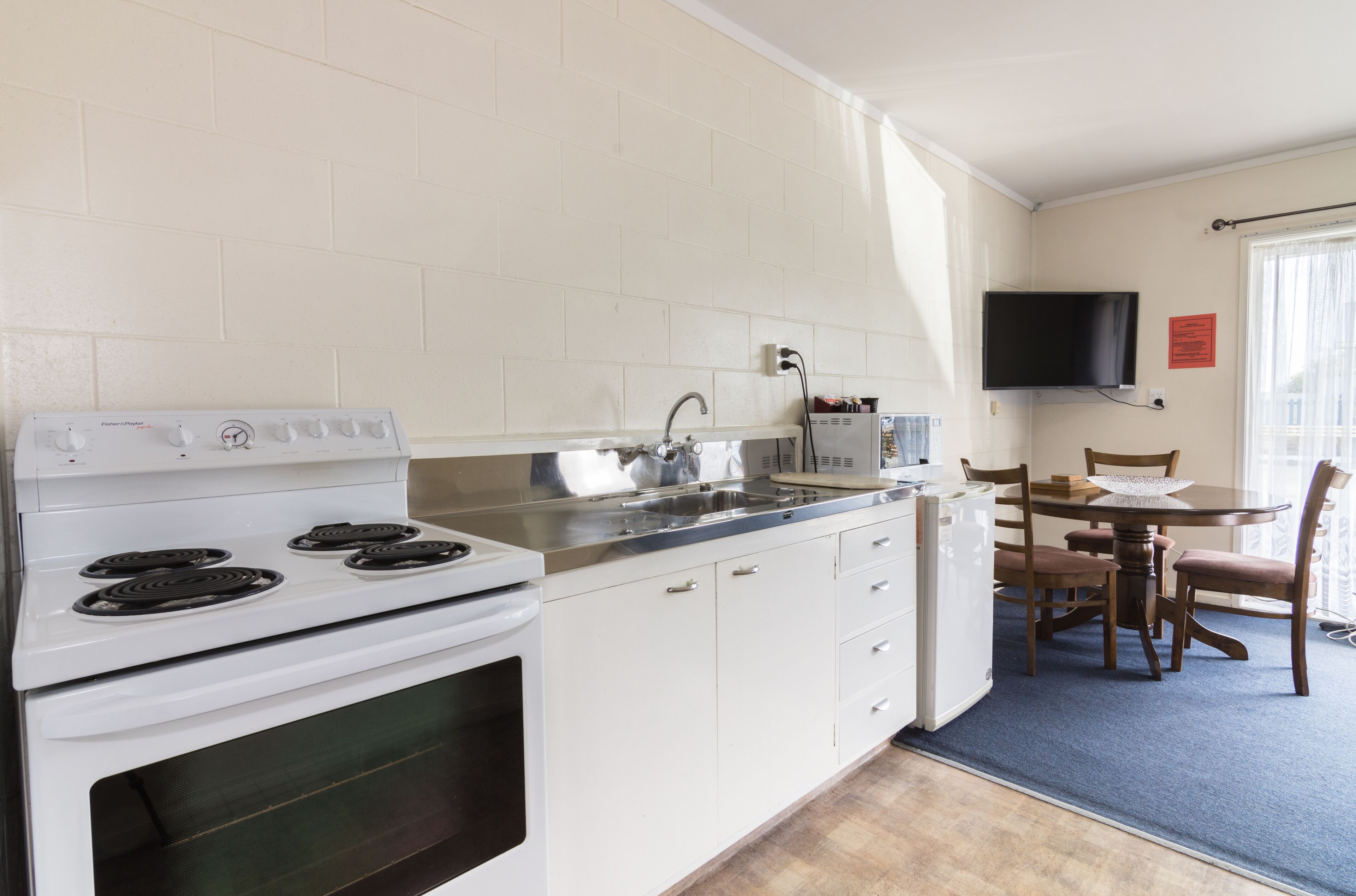 A compact kitchen with a stove, sink, and microwave. There is a dining table with chairs and a TV mounted on the wall.