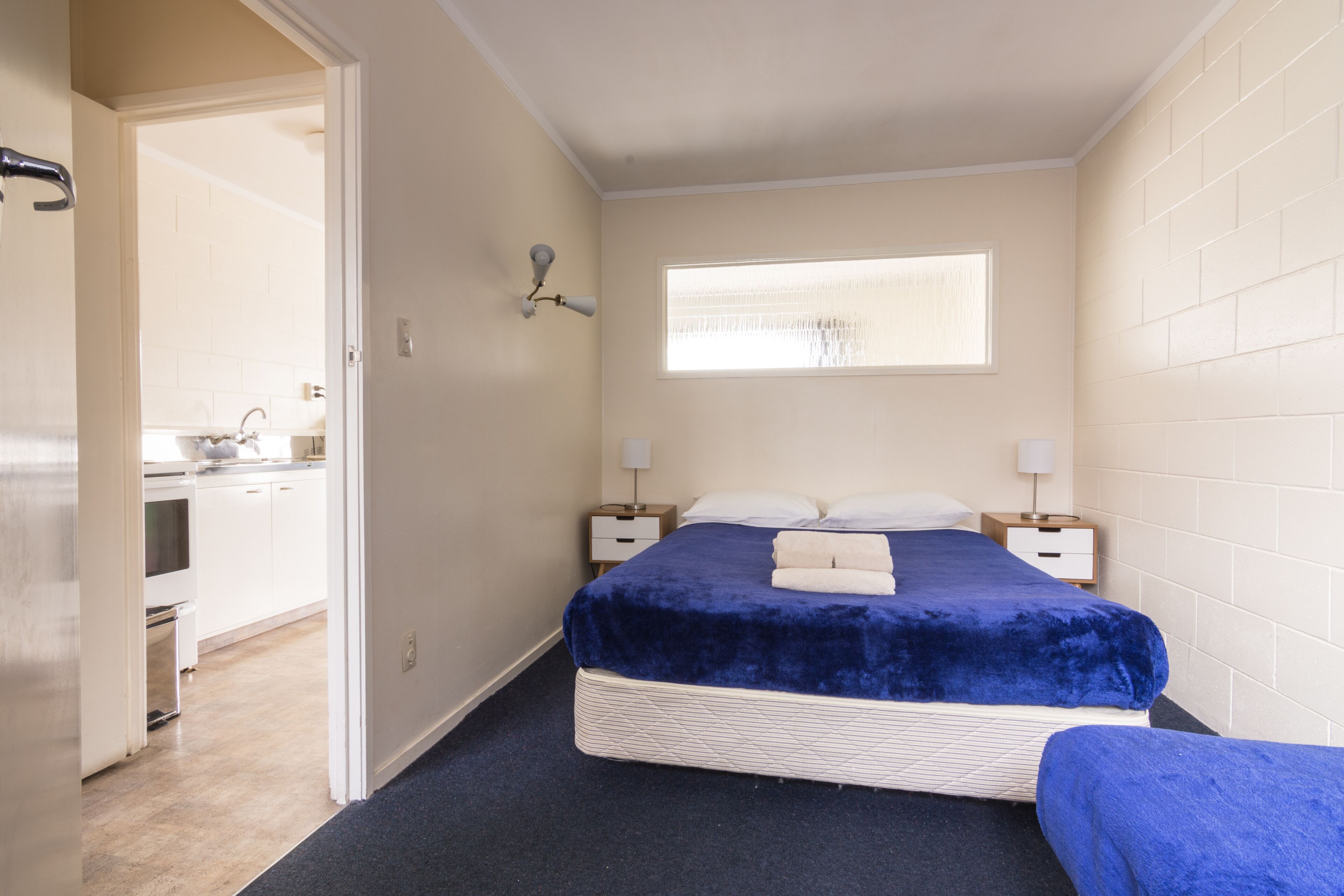 Ein Schlafzimmer mit einem Einzelbett, zwei Nachttischen, einem Fenster und Blick auf eine Küchenzeile.