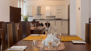 Un comedor con una mesa de madera dispuesta para cuatro personas, incluyendo platos, cubiertos y vasos. Hay sillas alrededor de la mesa, y se ve una cocina al fondo.