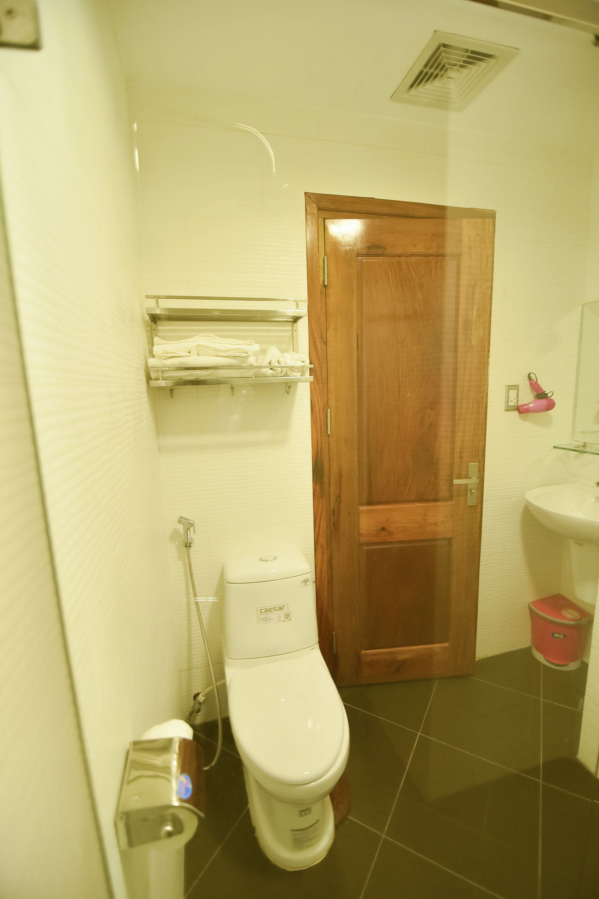 A bathroom with a toilet, a wooden door, a shelf with towels, a sink, and a shower.