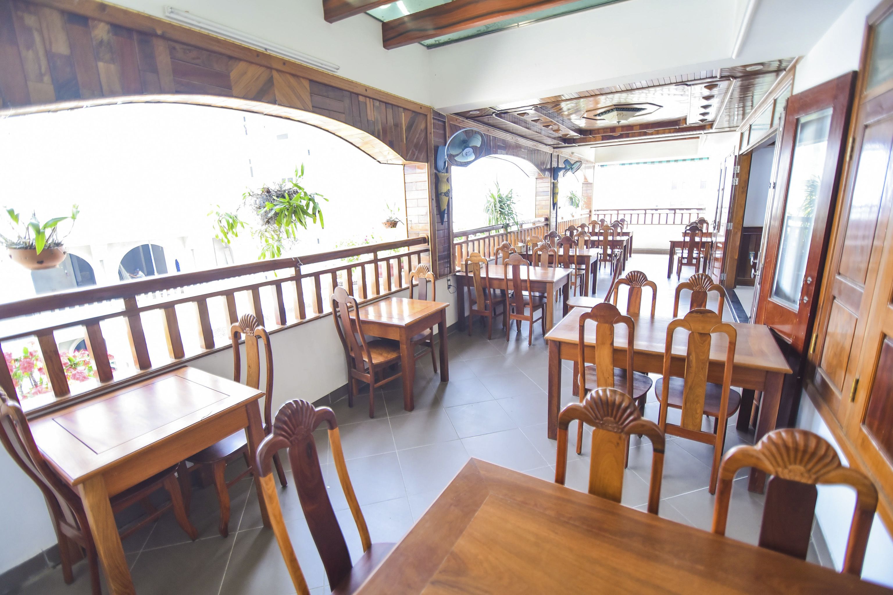 Un restaurant avec des tables et des chaises en bois, de grandes fenêtres ornées de plantes en pot et un balcon doté d’une balustrade.