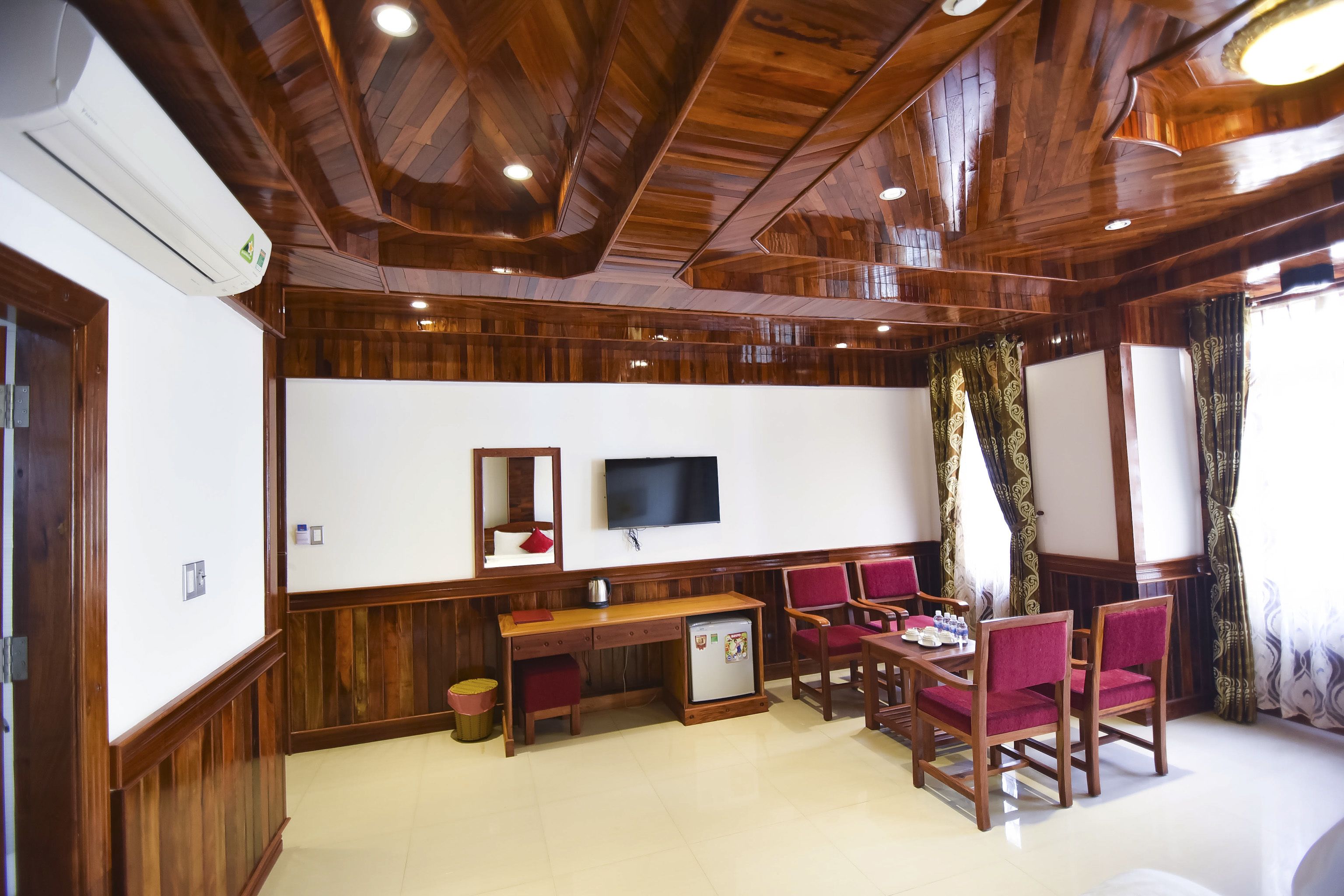 A room with wooden paneling, a flat-screen TV, a desk with chairs, and a small refrigerator.