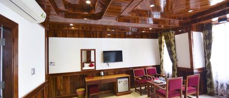 A room with wooden paneling, a flat-screen TV, a desk with chairs, and a small refrigerator.