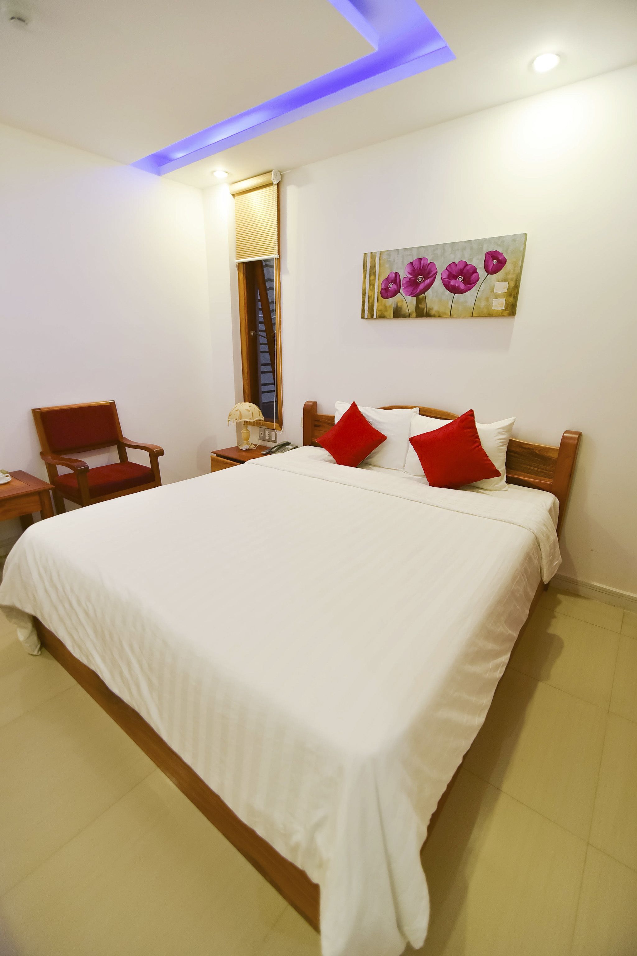 A neatly made bed with white bedding and red pillows, a wooden headboard, a wooden chair, a bedside table with a lamp, and a painting of flowers on the wall.