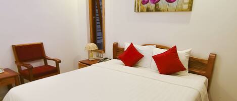A neatly made bed with white bedding and red pillows, a wooden headboard, a wooden chair, a bedside table with a lamp, and a painting of flowers on the wall.