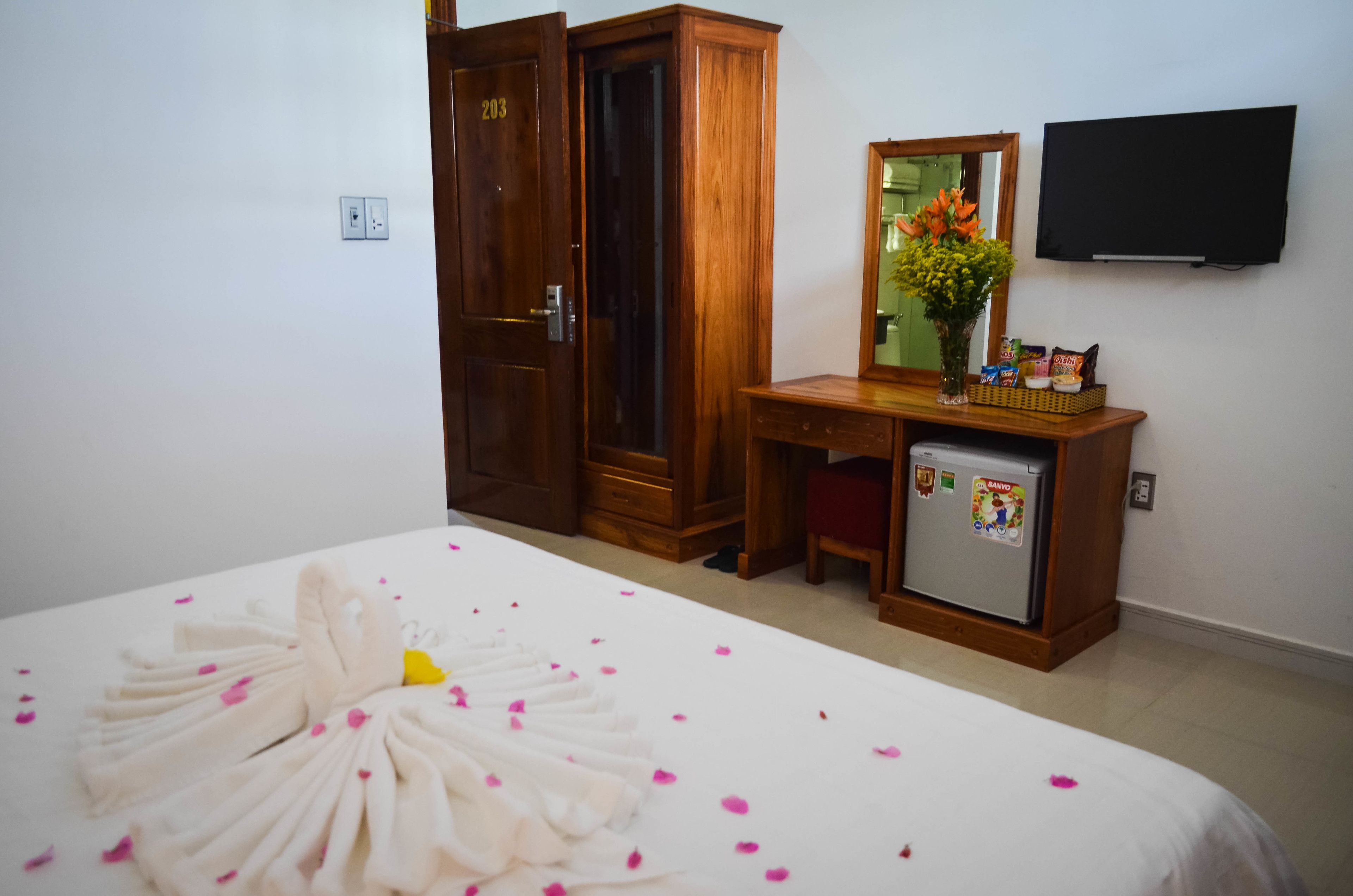 A hotel room with a bed, a wooden wardrobe, a desk with a mirror, a refrigerator, and a television on the wall.