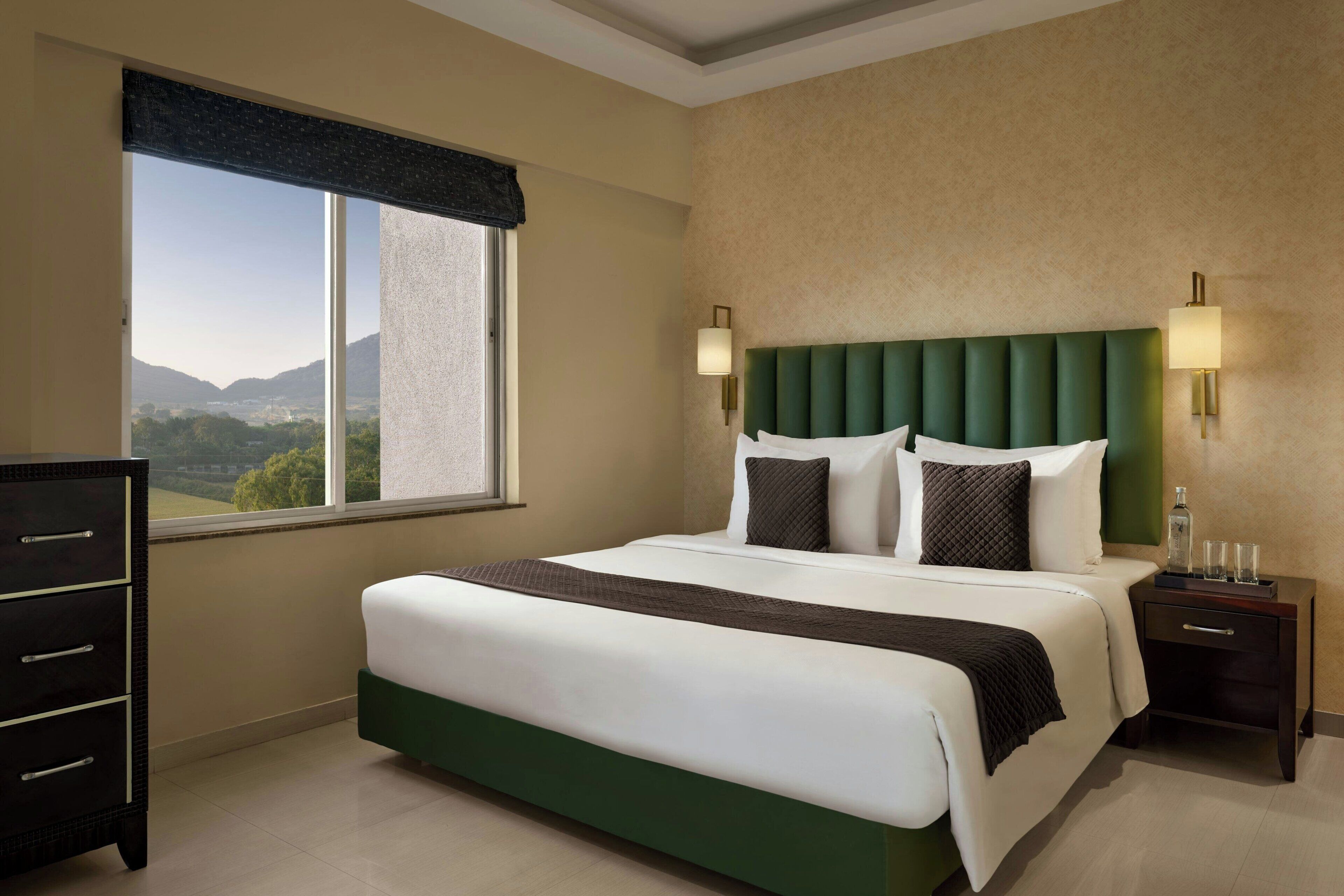 A hotel room with a large bed, a dresser, a nightstand with a lamp, and a window with a view of mountains.