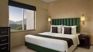 A hotel room with a large bed, a dresser, a nightstand with a lamp, and a window with a view of mountains.