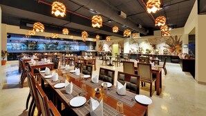 A spacious restaurant with multiple dining tables set for service, overhead pendant lighting, and a view of the ocean through large windows. - The Fern Residency Pune, Vadgaon-Talegaon, Series by Marriott (Mawal)
