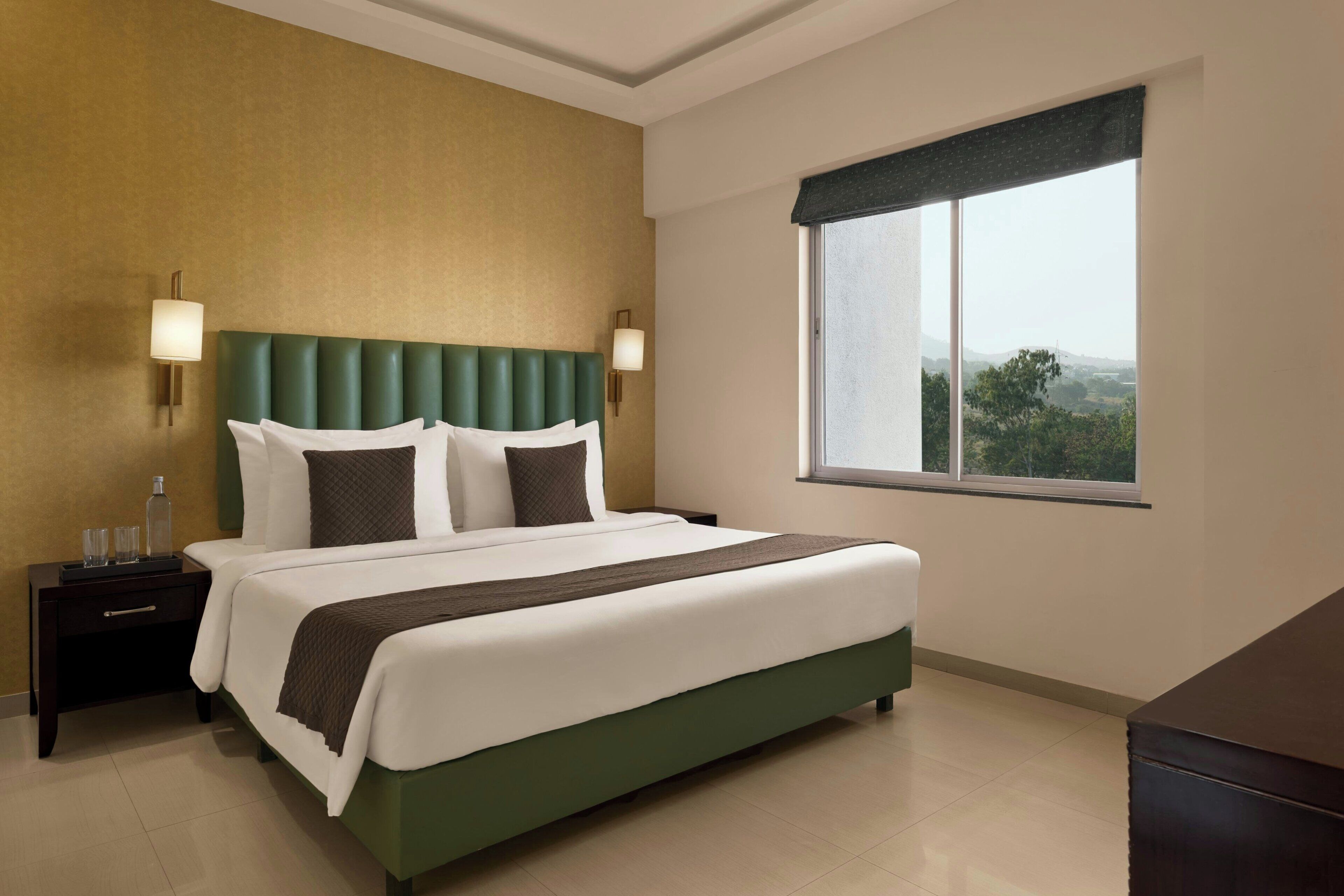 Una habitación de hotel con una cama grande, dos lámparas de noche, una mesita de noche, una ventana con vistas y un aparador.