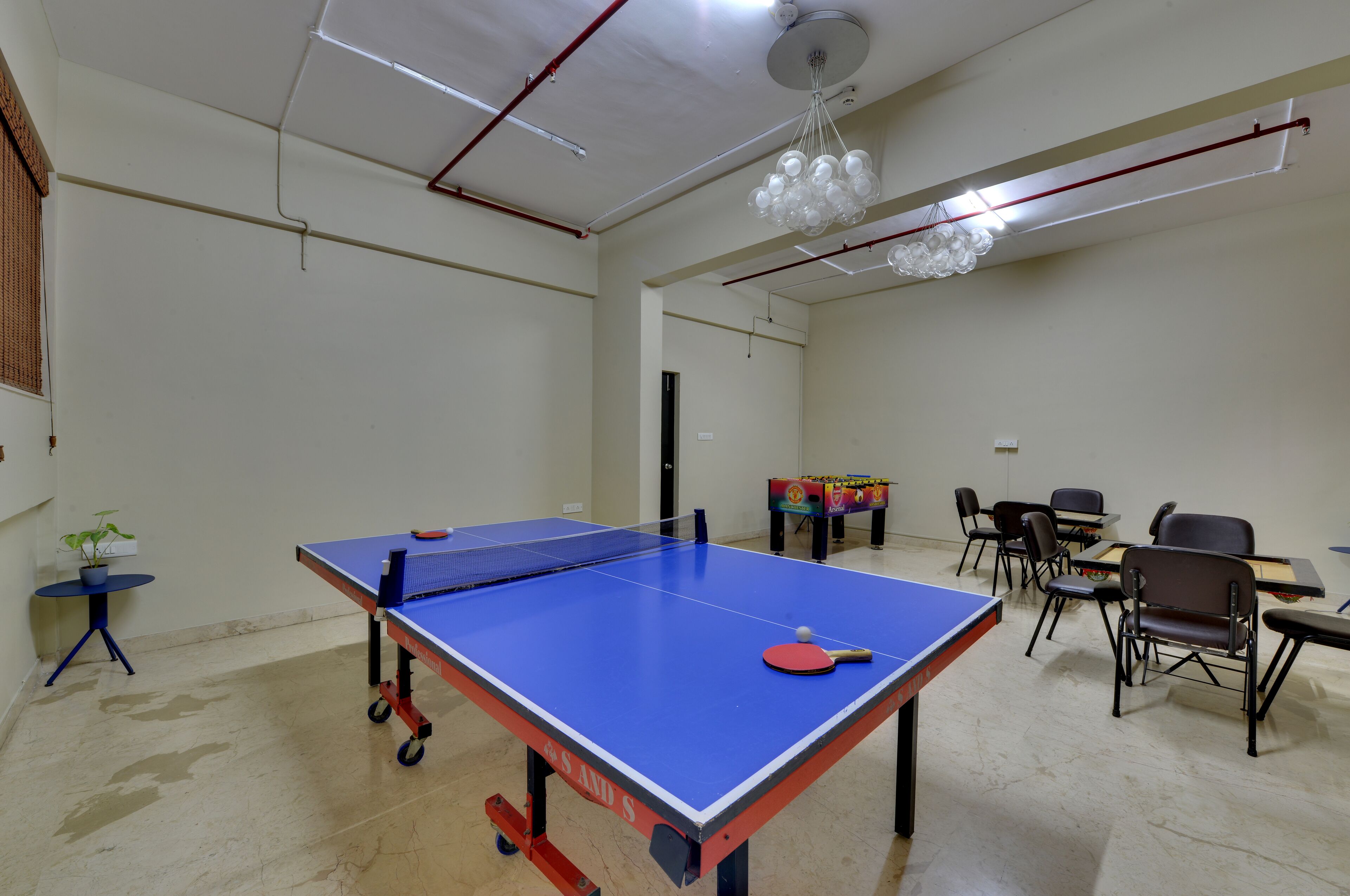 A ping pong table with chairs around it, a small table with a plant, and a table tennis table in a room with white walls and a ceiling light.