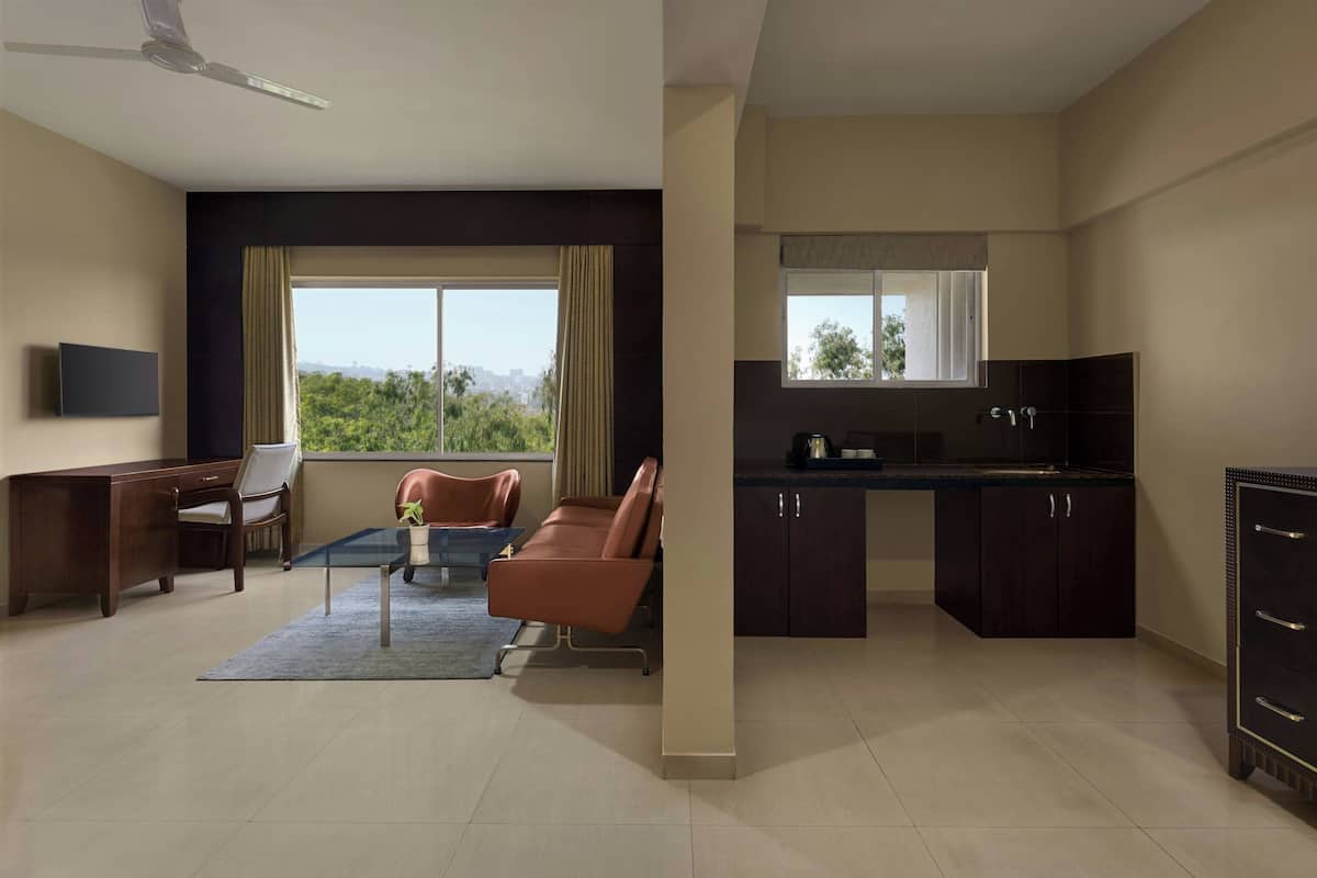 A modern hotel room with a flat-screen TV, a sofa, a coffee table, a desk, and a view of greenery.